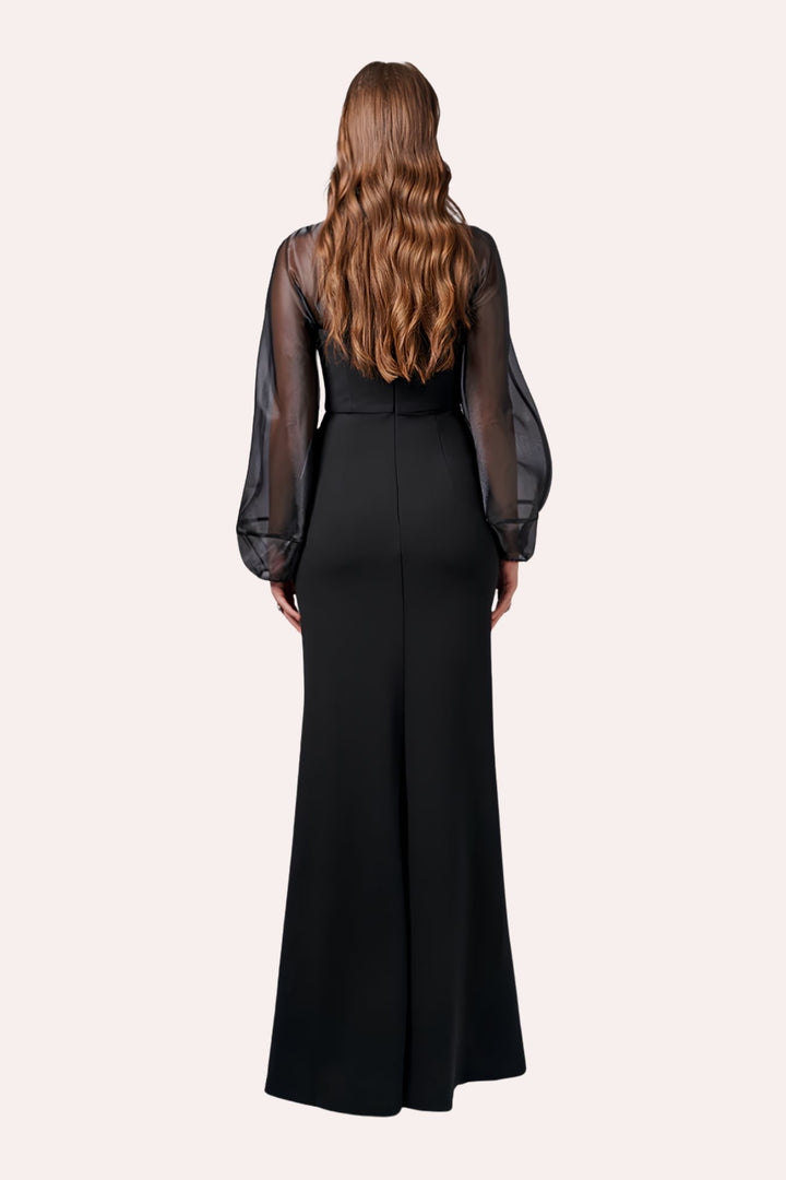 A woman viewed from the back wearing a long black dress with sheer mesh sleeves and a structured waist, against a plain light beige studio background.
