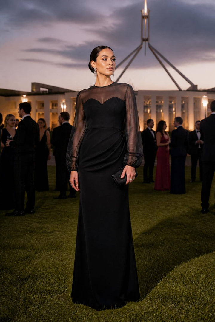 A woman wearing a floor-length black evening gown with sheer long sleeves and a sweetheart neckline stands outdoors on a lawn at dusk, with guests and a lit building in the background.