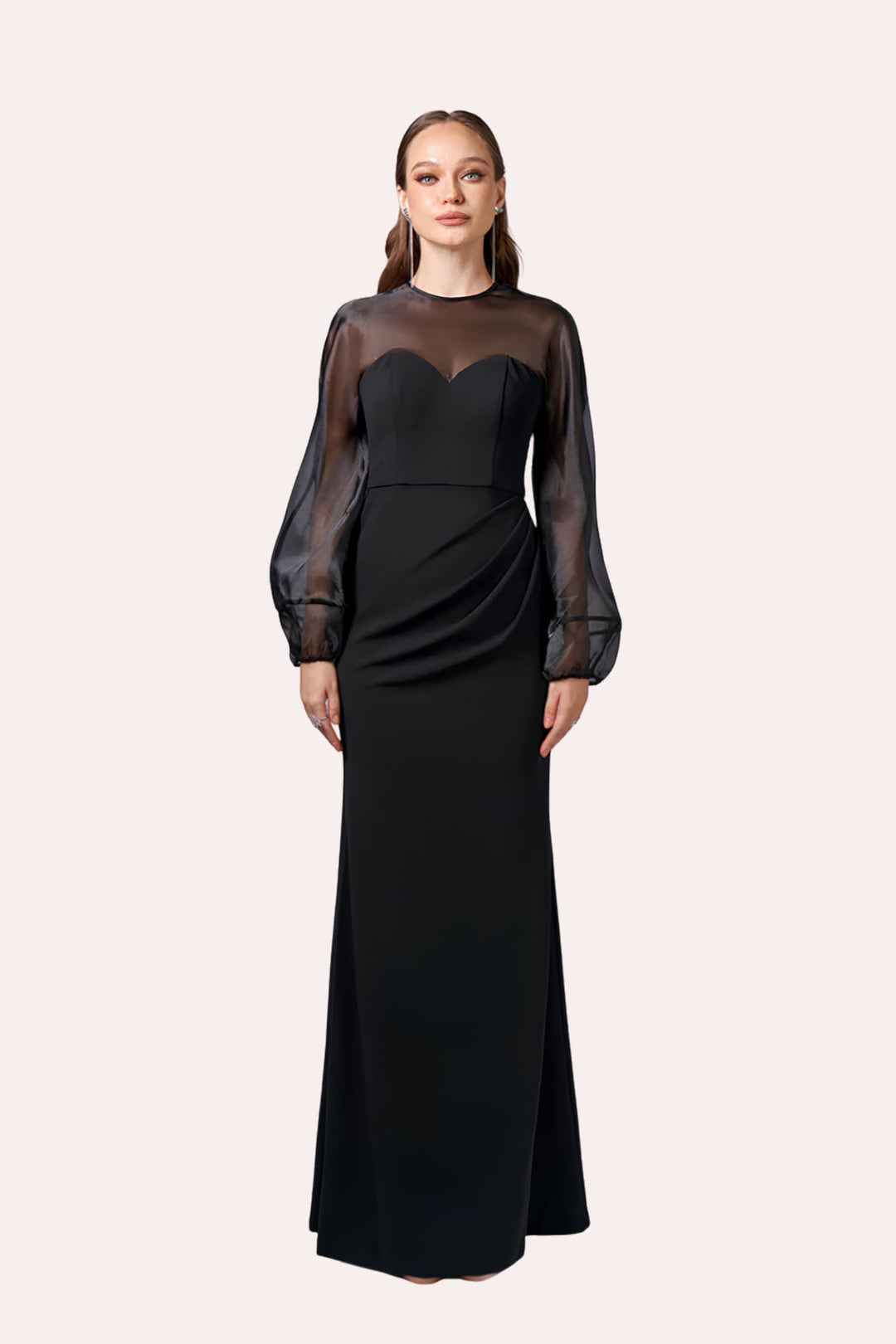 A woman modeling a floor-length black gown with sheer long sleeves and a sweetheart neckline, photographed against a plain light beige studio background.