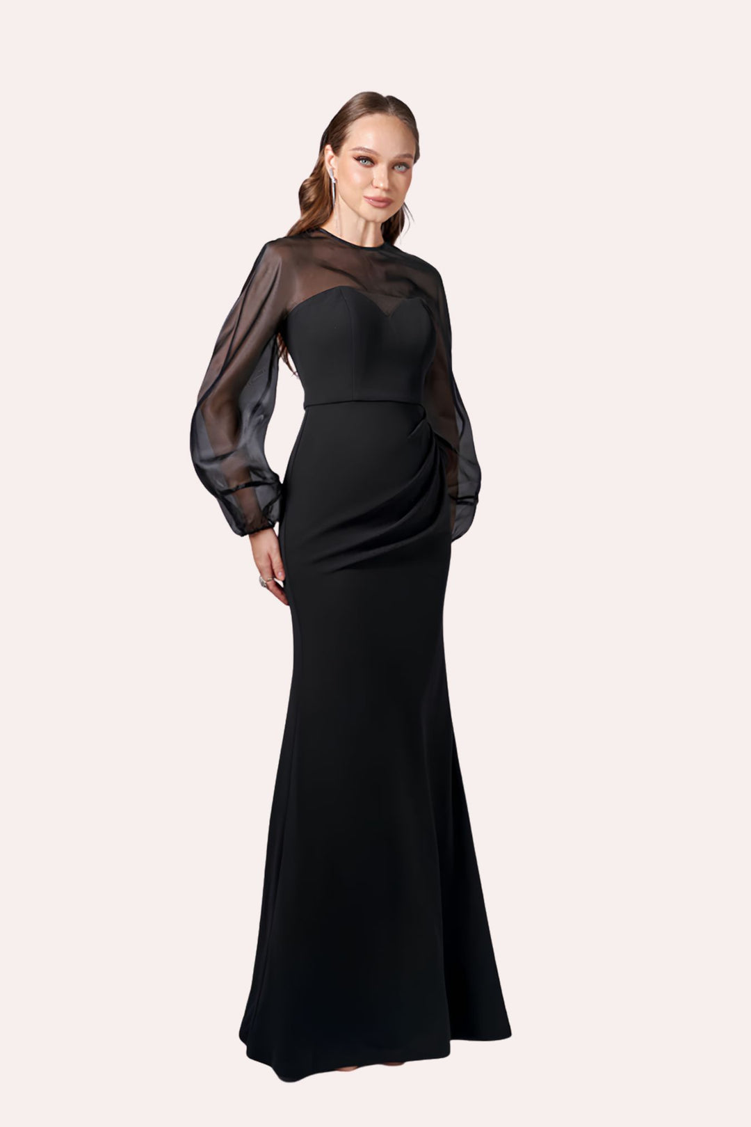 A woman modeling a floor-length black gown with sheer long balloon sleeves and a structured bodice, photographed against a plain light warm-gray studio background.