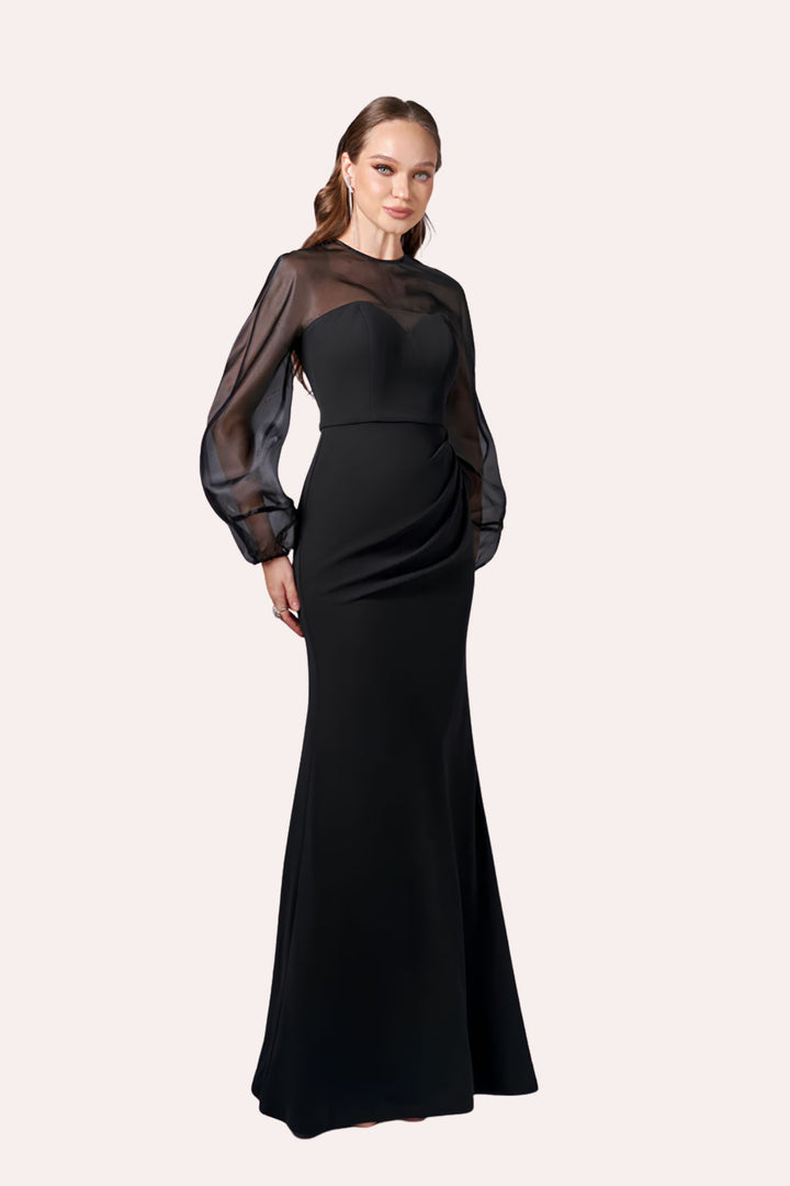A woman modeling a floor-length black gown with sheer long balloon sleeves and a structured bodice, photographed against a plain light warm-gray studio background.