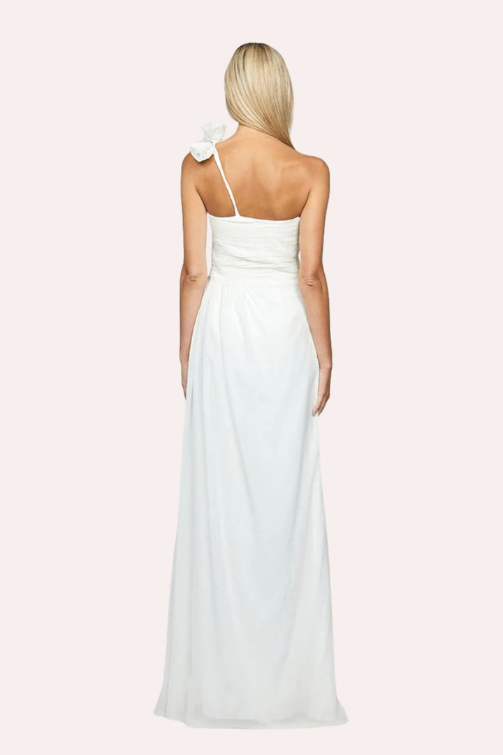 A woman shown from the back wearing a white sleeveless maxi dress with thin shoulder straps, a bow detail on one shoulder, and a gathered waist, against a pale pink studio background.