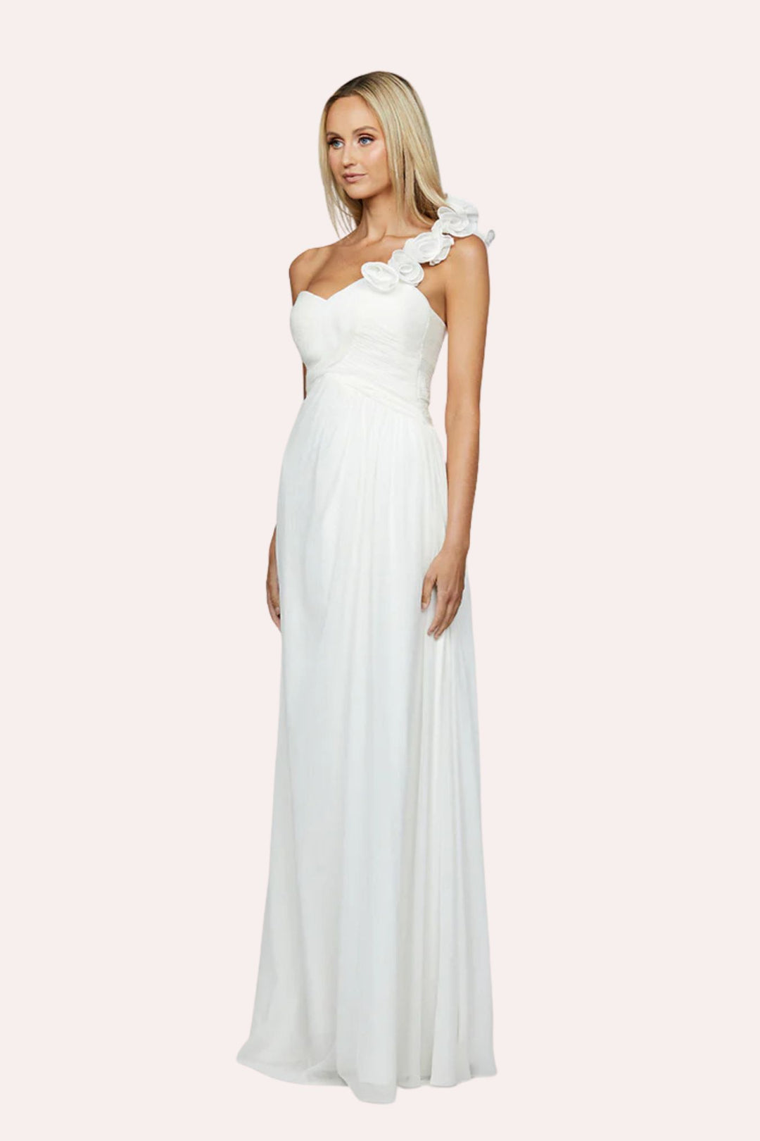 A blonde model wearing a floor-length white one-shoulder gown with a large fabric flower detail on the shoulder, photographed against a solid pale pink background.