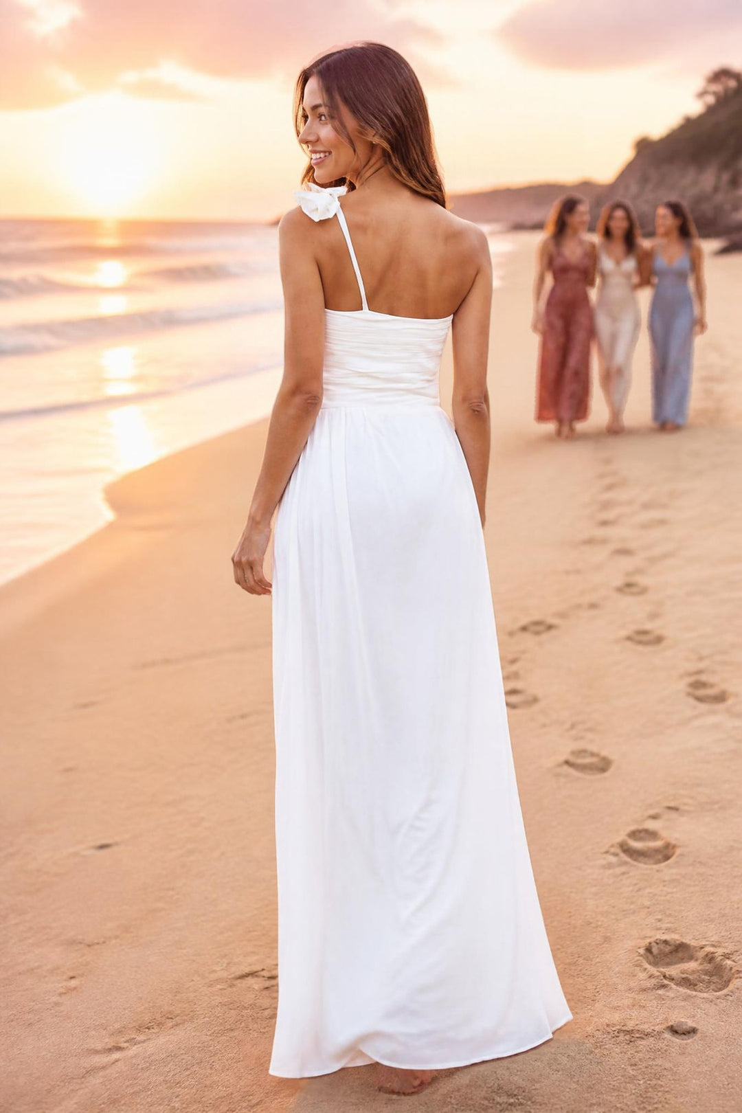 A woman stands on a sandy beach at sunset wearing a long white maxi dress with a single thin strap and a large bow detail at the neckline. Three other women are blurred in the background near the water.