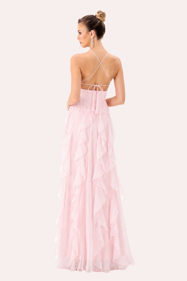 A woman shown from the back wearing a long light pink tulle dress with thin crisscross straps and a tie at the back, photographed against a plain light warm-gray studio background.