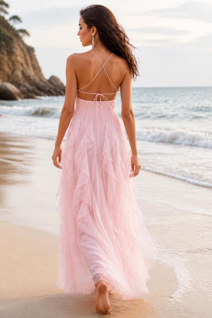 A woman wearing a long, flowing light pink tulle dress walks barefoot on a sandy beach with the ocean and rocky cliffs in the background.