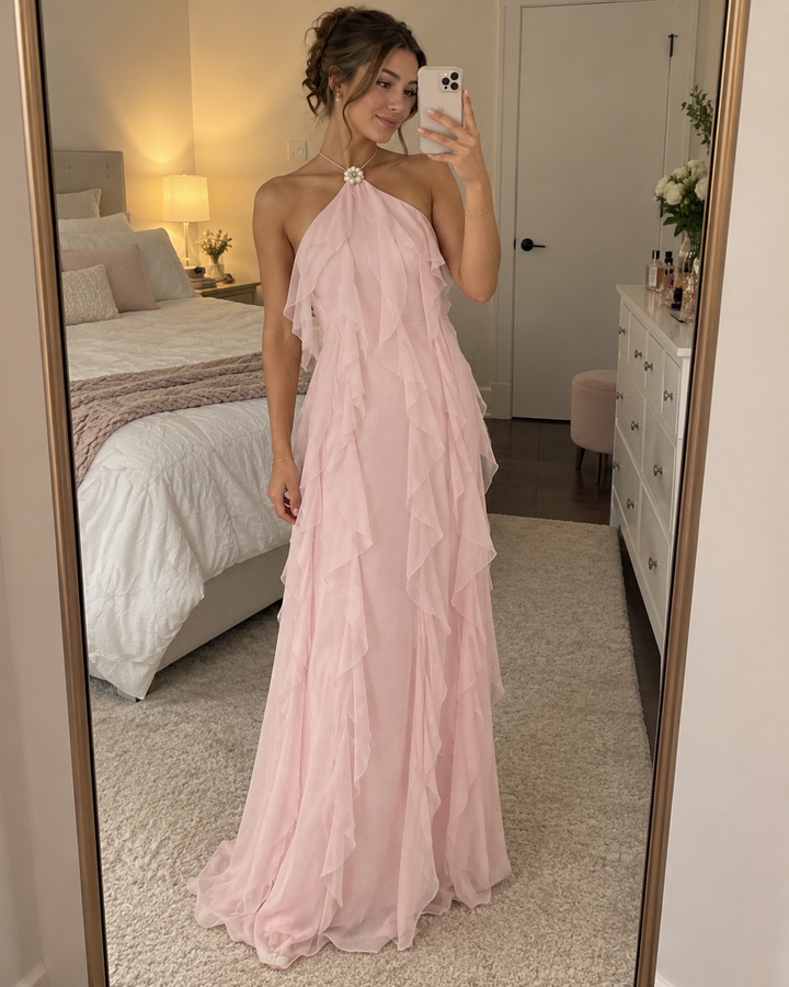 A woman takes a mirror selfie in a bedroom wearing a long, light pink halter gown with layered ruffle details. The bedroom features a bed with white bedding, a nightstand with a lamp, and a white dresser.